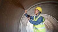 A female engineer in a hardhat and safety vest uses a hardness tester to test steel inside the steel pipe